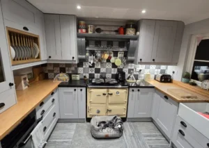 Kitchen featuring light grey cabinets, wooden countertops, a ceiling with two recessed lights, and a cream-colored stove surrounded by kitchen utensils, appliances, and decorative items. A pet bed is on the floor.