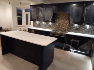 Kitchen with dark cabinets, white worktops, mosaic tile backsplash, central island, under-cabinet LED lighting, gas cooker, and ceiling spotlights.