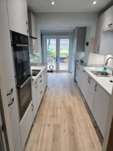 Galley kitchen with white cabinets, white worktops, built-in black appliances, vinyl flooring, and French doors opening to a garden.