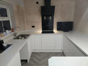 L-shaped kitchen with white cabinets, white countertops, a black glass induction hob and hood, grey herringbone flooring, and freshly plastered walls.