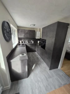 U-shaped kitchen with dark grey cabinets, a window providing natural light, minimalist decor, a wall clock, sink, boiler, and clean countertops.