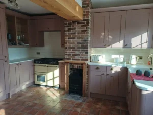 Country-style kitchen with pale pink cabinets, white countertop, vintage stove, fireplace, brick accent wall, tiled floor, vintage appliances, and sunlight streaming through a window.