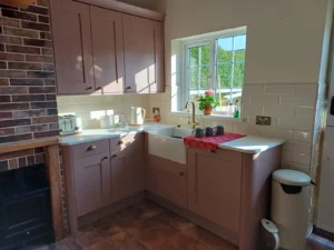 Country-style kitchen with pale pink cabinets, white countertop, a window with a view of greenery, and rustic charm featuring a brick wall and vintage appliances.