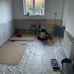 Renovation in progress: Room with bare walls, cardboard-covered flooring, a vacuum cleaner, tools, and a radiator visible.