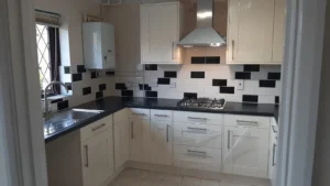 Newly renovated kitchen with a gas stove, black and white tiled backsplash, black countertop, boiler, stainless steel hood and sink, and white cabinets. Natural light enters through the windows.