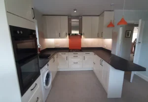 Kitchen featuring white cabinets, black worktops, an orange accent wall, cooker hood, oven, washing machine, and pendant lights.