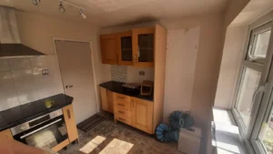 Kitchen with wooden cabinets, black countertop, oven, cooker hood, green mug, door, windows, ceiling spotlights, and blue ventilation ducts in the corner, illuminated by natural light.