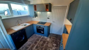 Kitchen with blue cabinets, stainless steel oven, washing machine, wooden countertop, doorway, and a sink beneath a window.