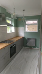 Kitchen with pale green walls, wooden countertop, grey cabinets, cooker hood, oven, white tiled backsplash, pendant lights, radiator, and windows.