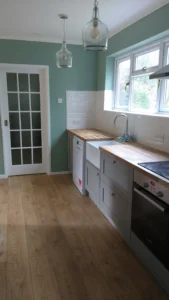 Kitchen with pale green walls, wooden floor, wooden countertop, silver appliances, grey cabinets, pendant lights, a glass door, and windows.