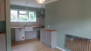 Kitchen with pale green walls, wooden countertop and floor, white dishwasher, radiator, cooker hood, pendant lights, and windows overlooking greenery.