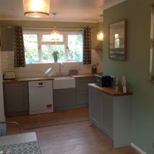 Kitchen with pale green wall and wooden floor, grey cabinets, farmhouse sink, wooden countertop, stylish curtains, pendant lighting, and windows with a view of greenery.