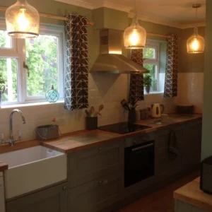 Modern kitchen with grey cabinets, farmhouse sink, wooden countertop, cooker hood, oven, stylish curtains, warm pendant lights, and windows overlooking greenery.