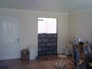Partially constructed wall and closed doorway in a room with wooden flooring, household clutter, and a ceiling bulb with holder attached.
