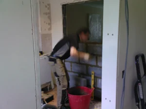 Construction worker applying mortar to a brick wall in a partially renovated room, with tools and materials visible.