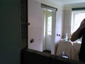 Partially renovated room with exposed walls, door to the outside, construction tools on a covered table, and a partially visible person wearing gloves.