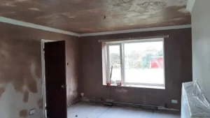 Partially renovated room with skimmed walls and ceiling, a large window, wall sockets, and an open door. Cups and a water bottle sit on the windowsill. A broom leans against the wall, and renovation debris is visible on the floor.