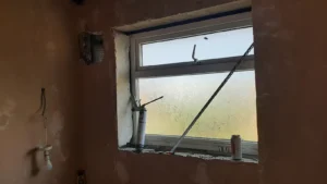 Renovation in progress: foggy window with a caulking gun and soda can on the windowsill, and a bulb with holder hanging from the wall.