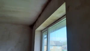 Partial view of a room's upper corner with plastered walls and a large window offering an outside view.