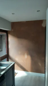 Partial view of a kitchen with skimmed wall, stainless steel sink, partially visible window, white ceiling with recessed lights, and white wooden floor. Natural light casts soft shadows.