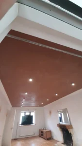 Room with brown ceiling and recessed lighting, wooden floor, flat-screen TV, wooden entertainment unit, radiator, broom, an open door, and fireplace with a mirror above it.