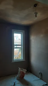 Renovation in progress: bare, unpainted room with window, ceiling light, skimmed ceiling and walls, and a bag of materials on the floor.